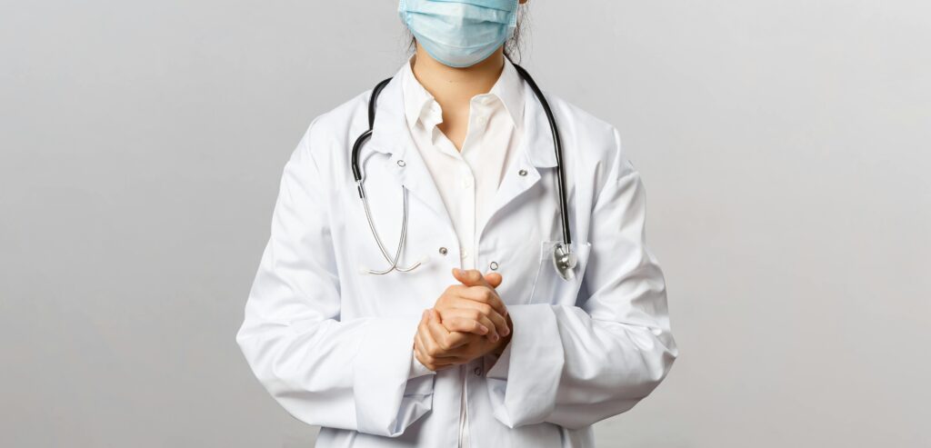 Healthcare professional wearing a mask and lab coat with stethoscope in neutral background.